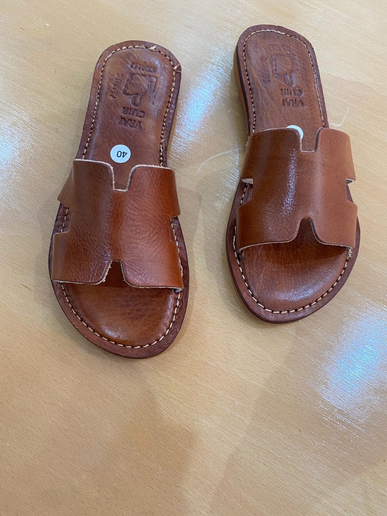 "A pair of artisan handcrafted Moroccan leather flat sandals featuring a classic -style strap. Made from high-quality natural grain leather with meticulous hand-stitching, shown from a top-down view on a neutral surface."