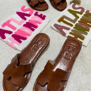 Peut inclure: Deux paires de sandales plates en cuir marron avec une bride en forme de H. Chaque paire est accompagn&eacute;e d'une pochette en tissu blanc avec le texte brod&eacute; ATLAS HANDMADE en fils rose et magenta ou olive et marron.