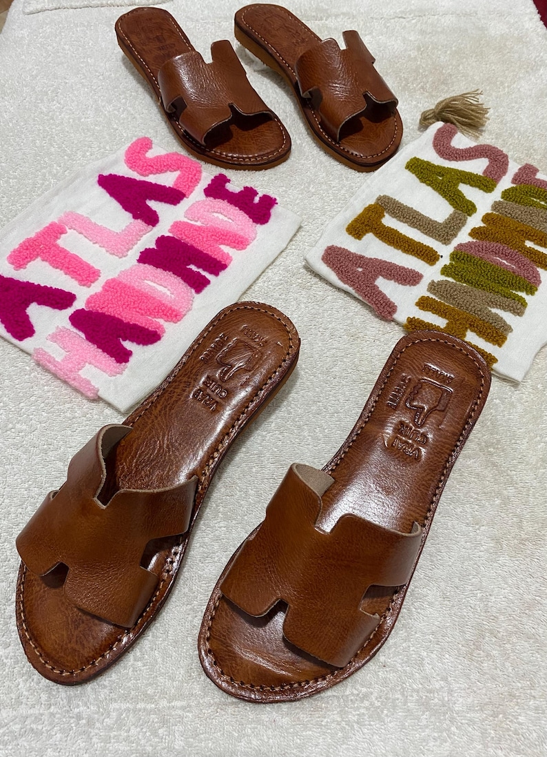 Peut inclure: Deux paires de sandales plates en cuir marron avec une bride en forme de H. Chaque paire est accompagn&eacute;e d'une pochette en tissu blanc avec le texte brod&eacute; ATLAS HANDMADE en fils rose et magenta ou olive et marron.