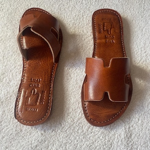 Peut inclure: Une paire de sandales plates en cuir marron avec une large bride en forme de H sur le dessus. La semelle int&eacute;rieure est estampill&eacute;e du texte VRAI CUIR, SKALLI et MAROC, accompagn&eacute; d'une ic&ocirc;ne repr&eacute;sentant une main pointant vers le haut.