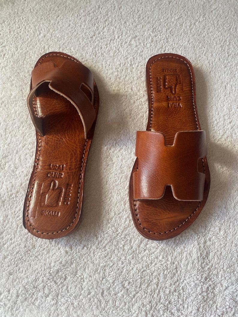 Peut inclure: Une paire de sandales plates en cuir marron avec une large bride en forme de H sur le dessus. La semelle int&eacute;rieure est estampill&eacute;e du texte VRAI CUIR, SKALLI et MAROC, accompagn&eacute; d'une ic&ocirc;ne repr&eacute;sentant une main pointant vers le haut.