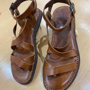 Könnte beinhalten: Ein Paar braune Ledersandalen mit mehreren Riemen und Knöchelschnallen. Die Sandalen haben eine genähte Sohle und eine dunkelbraune Gummibasis. Das Leder scheint abgenutzt zu sein, was auf einen Vintage- oder beliebten Artikel hindeutet.