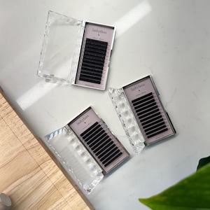 May include: Three open clear acrylic cases with black eyelash extensions. Each case has a pink interior with the "lashyfans" logo. The cases are on a white surface.