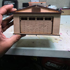 Puede incluir: Un garaje de madera en miniatura con un techo de lamas y una puerta de garaje con el logotipo de "Hot Wheels". La estructura es de madera marrón claro y tiene un diseño detallado, incluyendo una pequeña ventilación cerca del techo.