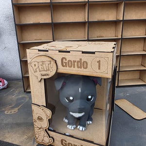 Puede incluir: Figura de perro blanco y negro dentro de una caja de madera. La caja está etiquetada como "PET!" y "Gordo 1", con el nombre "Gordo" repetido en la caja. La figura tiene patas blancas y una mancha blanca en la cara. La caja es de madera marrón claro.