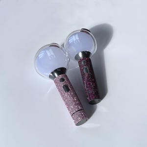 Op de afbeelding: Twee oplichtende concertstokken met doorzichtige bollen en met strass versierde handvatten. De stokken hebben de letters "BTS" op de bol gedrukt. Eén handvat is bedekt met roze strass, de andere met paarse strass. Op een witte ondergrond.