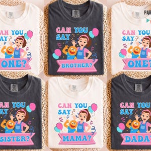 May include: Six folded t-shirts with colorful graphic designs. Each shirt features a cartoon character and the phrase "Can You Say" followed by a word like "One," "Brother," "Sister," "Mama," or "Dada." The shirts are in white and dark gray.