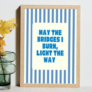 May include: A framed print with a light wood frame. The artwork features a cream-coloured rectangle with the text "MAY THE BRIDGES I BURN, LIGHT THE WAY" in blue. The background has blue vertical stripes.