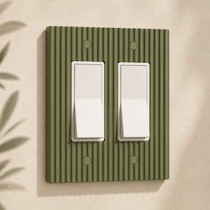 May include: A decorative olive green light switch cover with a ribbed texture. The cover has two white rocker light switches and four screw holes. The design is modern and minimalist.