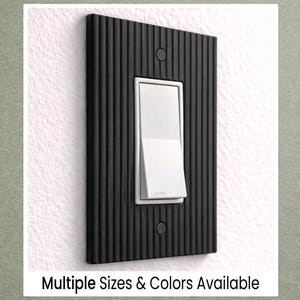 May include: A black, vertically-ribbed light switch plate with a white rocker switch. The plate is rectangular with two screw holes and is mounted on a white wall. Text at the bottom reads "Multiple Sizes & Colors Available."