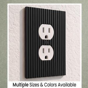 May include: Black, textured electrical outlet cover with two white outlets. The cover has vertical ridges and is mounted on a white wall. Text at the bottom reads "Multiple Sizes & Colors Available."