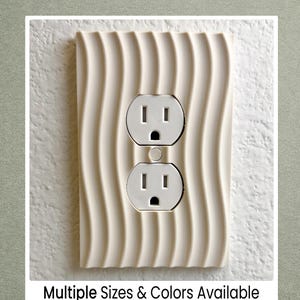 May include: A cream-colored, wavy-patterned outlet cover with two standard electrical outlets. The cover is mounted on a white wall. Text at the bottom reads "Multiple Sizes & Colors Available."