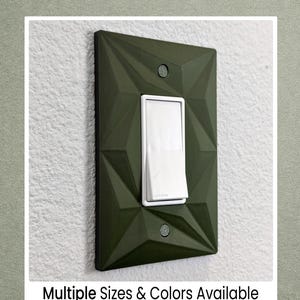 May include: A geometric olive green light switch cover with a white rocker switch. The cover has a textured, faceted design and is mounted on a white wall. The text "Multiple Sizes & Colors Available" is at the bottom.
