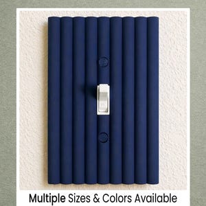 May include: A navy blue light switch cover with a ribbed design. The cover has a white toggle switch and two round screw holes. The text "Multiple Sizes & Colors Available" is at the bottom. The cover is mounted on a white wall.