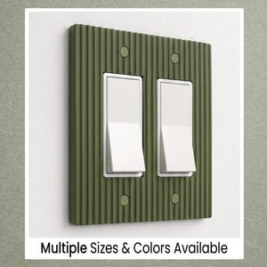 May include: Olive green double light switch cover with a textured, ribbed design. Features two white rocker switches and four small, round screws. The text "Multiple Sizes & Colors Available" is at the bottom.