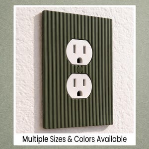 May include: Olive green textured electrical outlet cover with two white outlets. The cover has vertical ribbed detailing. The text "Multiple Sizes & Colors Available" is at the bottom of the image.