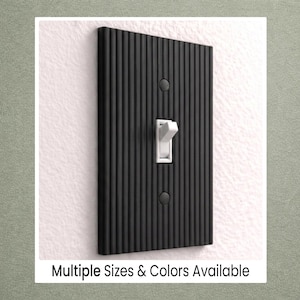 May include: A black, vertically-ribbed light switch cover with a white toggle switch. The cover is rectangular with two screw holes and is mounted on a white wall. Text at the bottom reads "Multiple Sizes & Colors Available."
