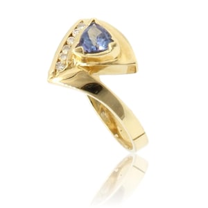 May include: A gold ring featuring a triangular-cut purple-blue gemstone set in a polished gold band. The band includes a curved section accented with five small, round, clear white diamonds arranged in a vertical row along the side of the setting.
