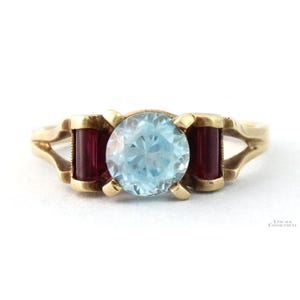 May include: A gold ring featuring a round, light blue gemstone center stone, flanked by two rectangular, dark red gemstones. The ring has a split shank design.
