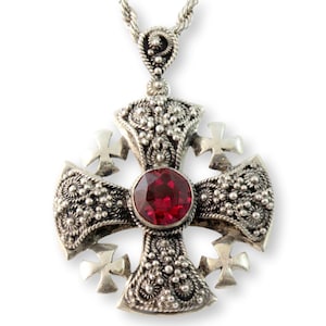 May include: Silver-toned pendant necklace featuring a detailed cross design. The cross has intricate filigree patterns, small crosses at the ends of each arm, and a large, round, red gemstone at the center. The chain is a twisted rope style.