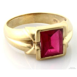 May include: A gold ring featuring a rectangular, faceted, deep pinkish-red gemstone set in a smooth, polished gold band. The interior of the band is clearly stamped with the text 10K, indicating the gold purity of the metal setting.