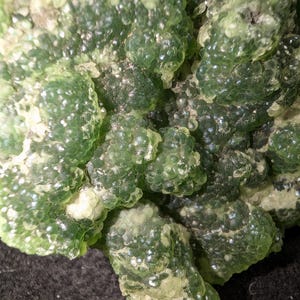 May include: Close-up of a mineral specimen featuring a cluster of small, translucent, green, bubble-like formations. The surface has a crystalline appearance with areas of lighter green and white. The overall texture is rough and uneven.