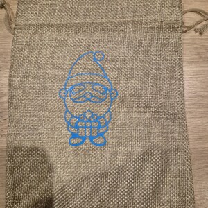 May include: A rectangular beige burlap drawstring bag featuring a bright blue graphic print of a stylized gnome wearing a hat and holding a small gift box. The bag has a woven texture and includes matching beige drawstrings at the top closure.