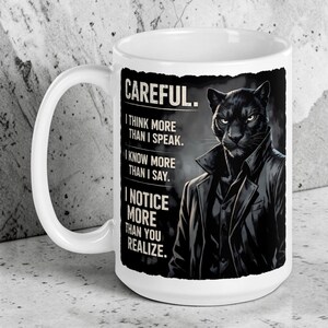May include: A white ceramic mug featuring a graphic of a black panther wearing a dark coat. The text reads: CAREFUL. I THINK MORE THAN I SPEAK. I KNOW MORE THAN I SAY. I NOTICE MORE THAN YOU REALIZE. The design is set against a dark, textured background.