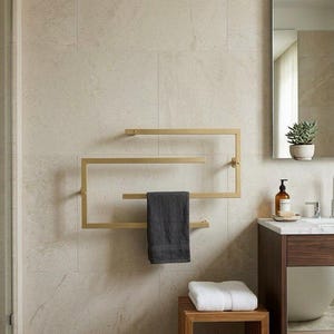 Può includere: Un portasciugamani geometrico color oro montato su una parete piastrellata beige. Un asciugamano grigio scuro è appeso al portasciugamani. Uno sgabello di legno contiene un asciugamano bianco piegato. Uno specchio e un mobile da bagno sono visibili.