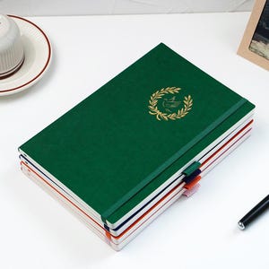 May include: A stack of four rectangular notebooks with elastic closures. The top notebook is dark green with a gold laurel wreath and bird emblem. The notebooks below are navy blue, orange, and light pink, each featuring a matching elastic pen loop.