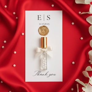 May include: A wedding favour featuring a small glass bottle filled with pearls, topped with a gold cap and a white bow. The bottle is on a white card with the initials "E|S", date "11.17.2025", a gold seal, and the words "Thank you". The background is red satin.