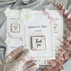 May include: White cards with floral accents and the text "Mahmoud & Safa" and "29.9.2025". Each card has a small square with "Thank you" written on it. White flower embellishments are attached to the cards.