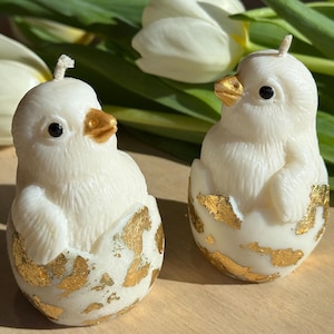 Puede incluir: Dos velas blancas con forma de pollito, con picos y detalles dorados, saliendo de mitades de cáscaras de huevo decoradas con pan de oro. Las velas están sobre una superficie de madera clara, con tulipanes blancos al fondo.