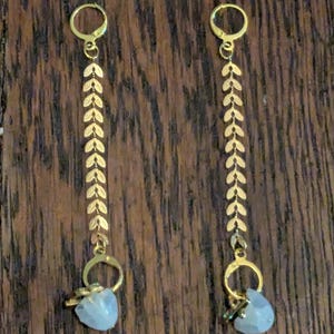 May include: A pair of gold-colored earrings. Each earring features a long chain of leaf-shaped charms, connected to a small, light blue gemstone. The earrings have a lever-back closure. The background is a dark wood surface.