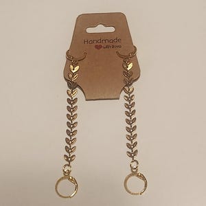 May include: Gold-colored dangle earrings with a leaf design, hanging from a brown tag that reads "Handmade with love." The earrings feature a series of small leaves along a chain, ending in a circular clasp.