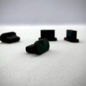 Könnte beinhalten: Vier kleine, schwarze, massive Kunststoffstopfen mit abgerundeter, leicht konischer Form. Jeder Stopfen hat eine flache, glatte Oberseite und ein geripptes, strukturiertes Seitenprofil, das zum Einsetzen in eine Öffnung vorgesehen ist.