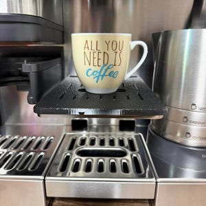 Könnte beinhalten: Eine beige Kaffeetasse mit dem Text "ALL YOU NEED IS coffee" in braunen und blauen Buchstaben steht auf einem schwarzen Tablett einer Espressomaschine aus Edelstahl. Die Maschine hat eine silberne Oberfläche und einen Wassertank.