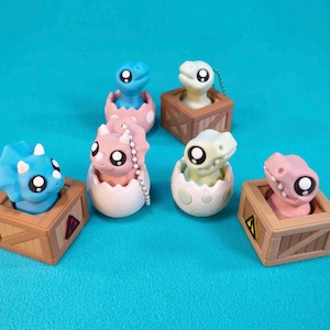 May include: A collection of colorful dinosaur figurines. The dinosaurs are in various colors, including blue, pink, and green. Some are in eggs, and others are in small wooden crates. The background is a solid teal color.