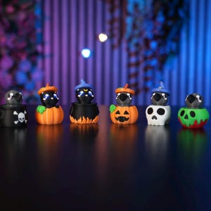 Puede incluir: Seis pequeñas figuras de gatos negros con ojos grandes, cada uno saliendo de un recipiente diferente: un caldero con calavera, una calabaza, un caldero con llamas, una calabaza de Halloween, una calavera blanca y una calavera verde.