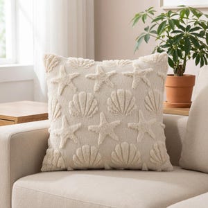 May include: A square throw cushion in a light cream colour featuring a raised, textured pattern of starfish and seashells across the front surface. The design is monochromatic, with the raised motifs created in the same shade as the base fabric.