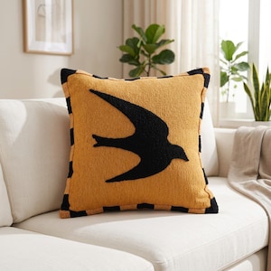 May include: Mustard yellow square cushion featuring a black bird design. The cushion has a textured surface and a black and tan chequered border. It is placed on a white sofa. The cushion measures approximately 40cm x 40cm.