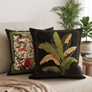 May include: Two square throw pillows. The left pillow features a cream background with a green border, displaying a colorful print of deer, birds, and flowers. The right pillow has a black background with a brown border and a large banana leaf design.