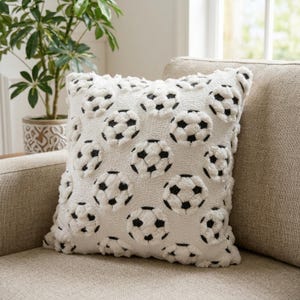 May include: A square, cream-coloured cushion featuring a repeating pattern of black and white footballs. The cushion is made of a textured fabric and is placed on a beige sofa. A potted plant is in the background.