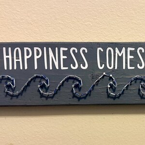 May include: A rectangular, dark gray wooden sign with the phrase "HAPPINESS COMES IN WAVES" in white lettering. Below the text, a wave design is crafted with silver wire. The sign is mounted on a light beige wall.
