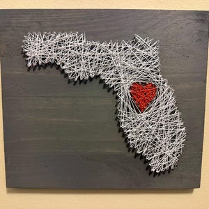 May include: A wooden string art piece depicting the state of Florida in white string, with a red heart in the center. The artwork is mounted on a dark gray stained wooden board, creating a striking contrast.