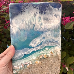 May include: Rectangular resin artwork showing an ocean scene. The top has a dark blue and white wave design, leading to turquoise and a sandy beach base with small shells. The piece is held against a backdrop of pink flowers.