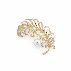 May include: A gold-coloured feather brooch, adorned with clear crystals and a hanging white pearl. The brooch features an intricate design with curved lines, set against a white backdrop. A piece of jewellery.