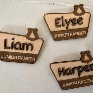 May include: Three trapezoidal wooden name badges featuring a dark brown shield icon with a pine tree cutout at the top. Each badge displays a name in raised dark brown lettering and the text JUNIOR RANGER in a lighter wood tone.