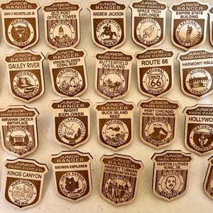 May include: A collection of twenty-eight wooden Junior Ranger badges in a shield shape. Each badge features engraved text and illustrations representing various national parks, landmarks, and historical sites, including Route 66 and various monuments.