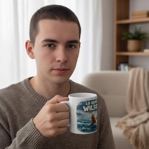Pode incluir: Caneca de cerâmica branca com uma ilustração de um galo num mar tempestuoso e o texto "LO SIENTO WILSO". A caneca tem uma pega branca. A pessoa que segura a caneca está a usar um camisola cinzenta.